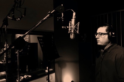 Male wearing glasses with headphones on in front of a microphone in a recording studio.